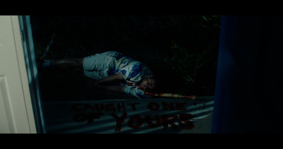 A bloodied man is lying on the floor with his arms splayed out. A message written in blood on the floor reads 'CAUGHT ONE OF YOURS